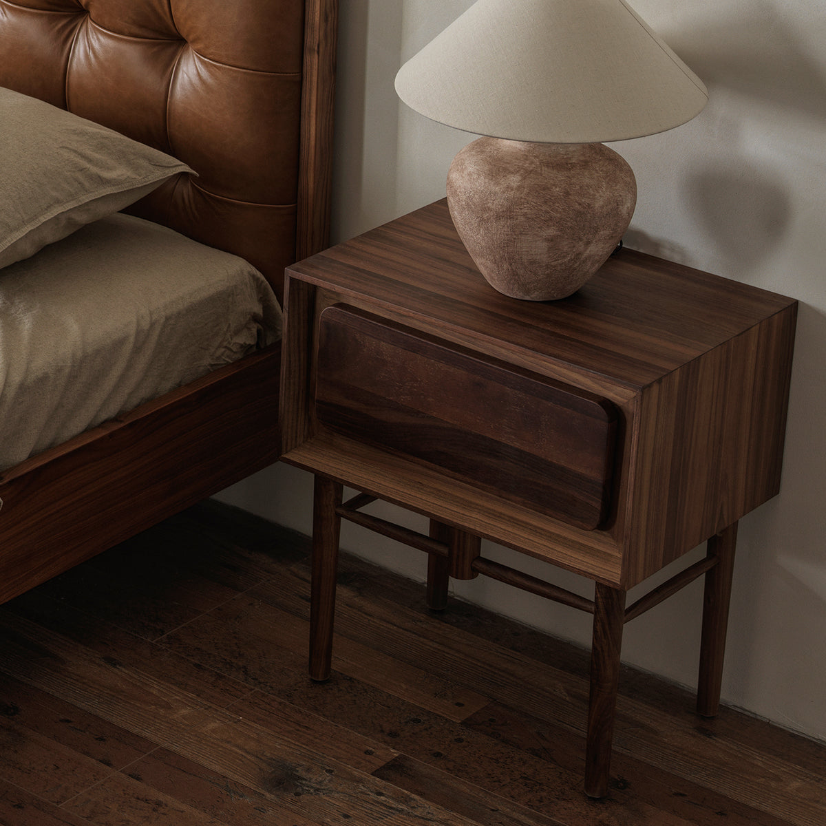 Three-quarter elevated view of walnut bedside table with table lamp beside a leather upholstered bed