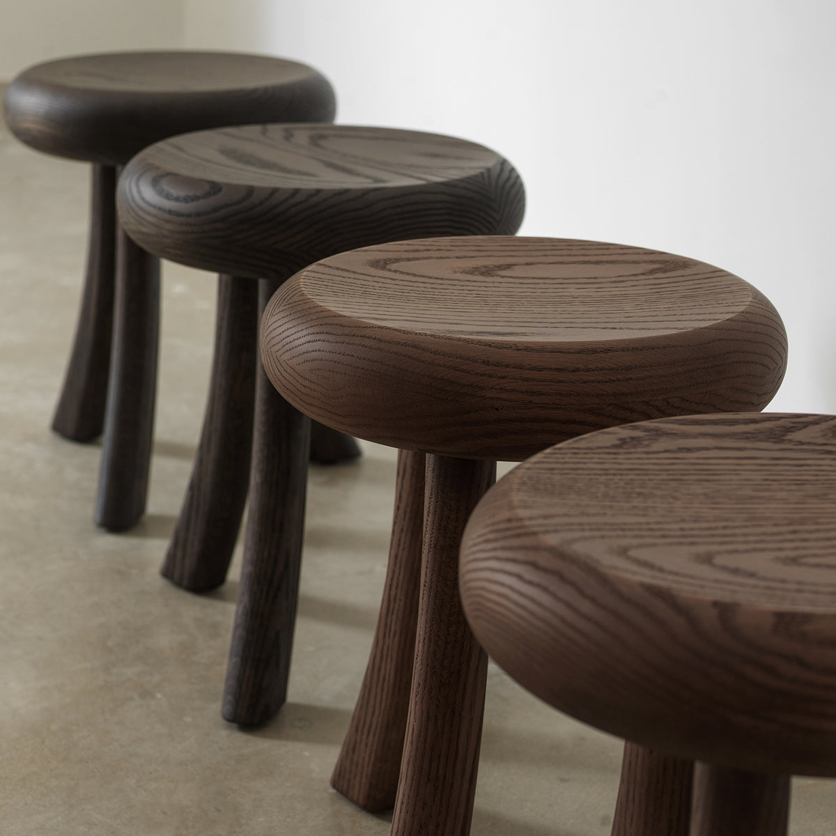 Angled perspective view of four solid wood 3-legged side tables lined up in various finish tones